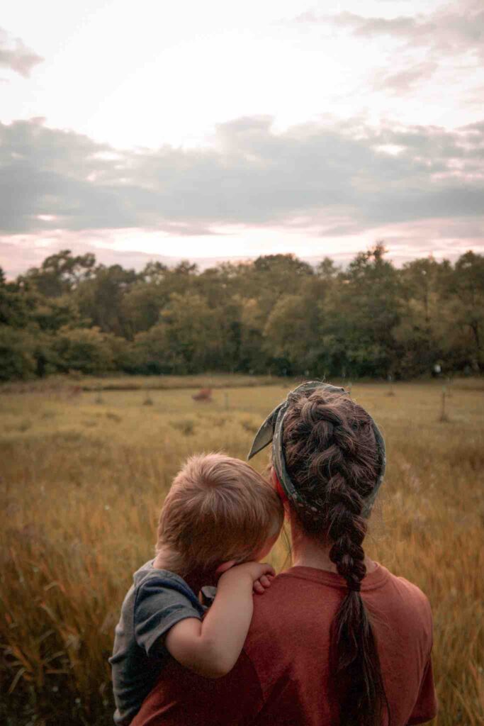 While hiking with toddler, mom is holding boy who rests head on her shoulder while looking out at deer in field.