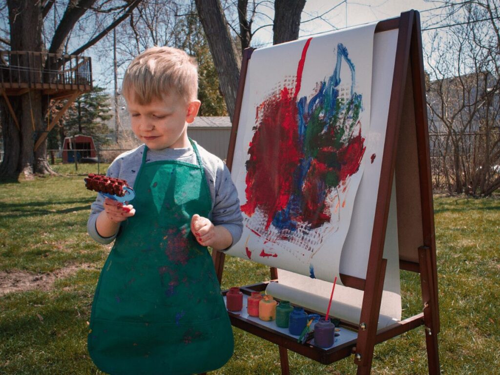 Outdoor creative arts ideas for kids: small boy painting on an easel in his backyard.