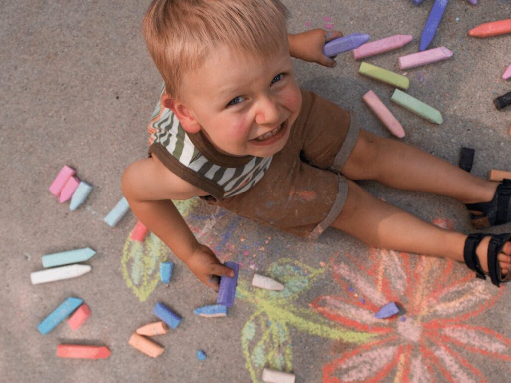 Small boy smiling up at camera while doing sidewalk chalk- summer bucket list