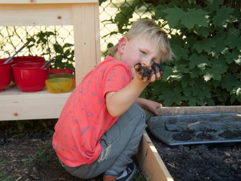 small boy holds up mud in his hand while playing in mud kitchen