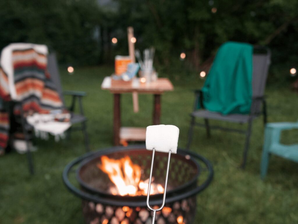marshmallow in focus in front of a campfire with chairs, blankets, and twinkle lights in the backgrount.