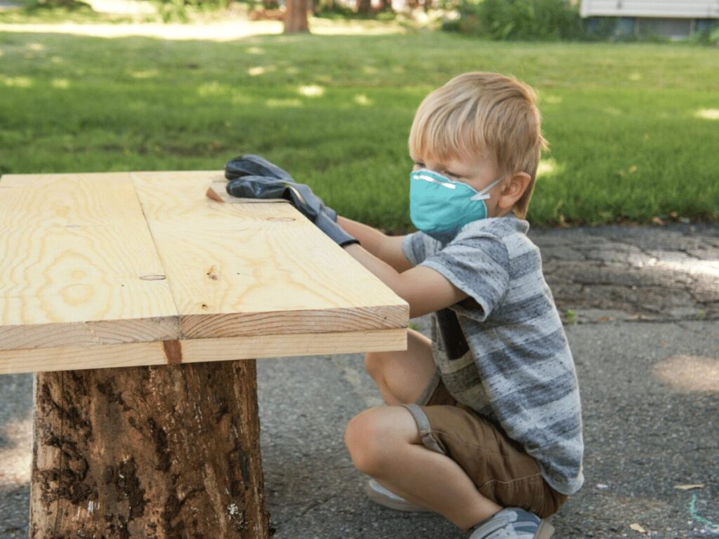 small boy wearing mask and gloves while sanding table top as part of furniture making business- business ideas for kids