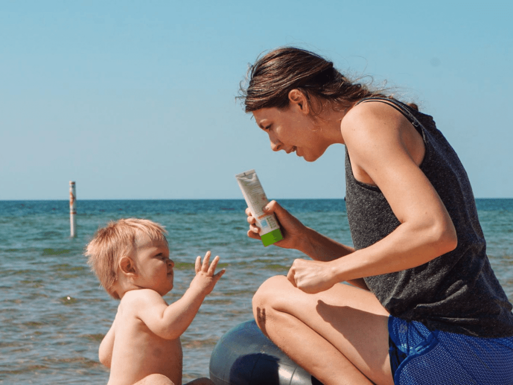 small boy and mom sitting on beach while mom holds up sunscreen- outdoor gear for babies