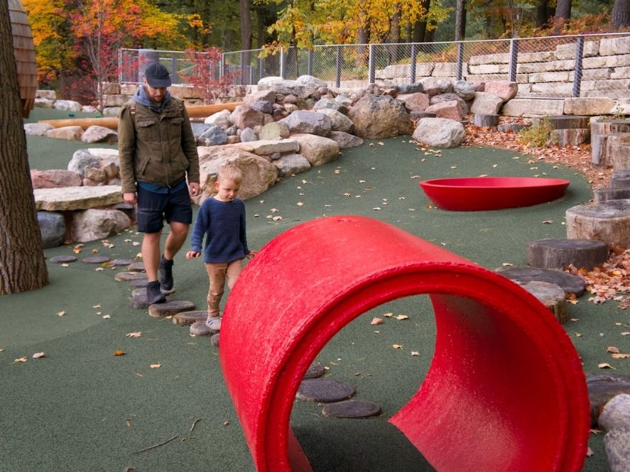 small boy and father walking on tree stumps toward large red tunnel on playground at dow gardens