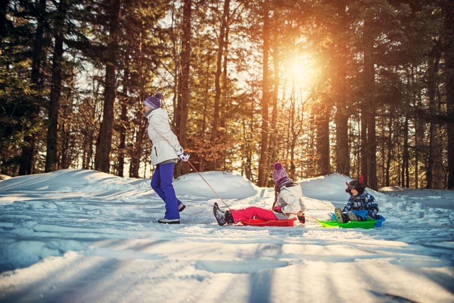 Mom pulling two kids on sleds through the woods while winter hiking with kids