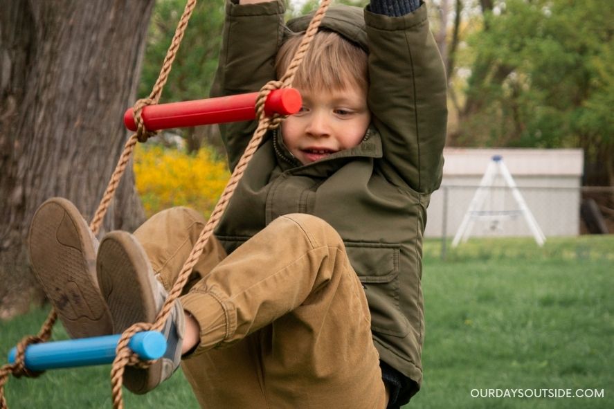 Young boy swinging from rope ladder in tree- improved gross motor skills are one of the benefits of outdoor play