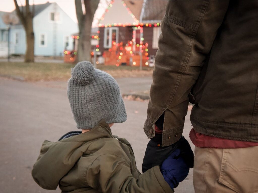 While on a Christmas scavenger hunt, small boy holds father's hand while looking at house decorated with Christmas lights. 
